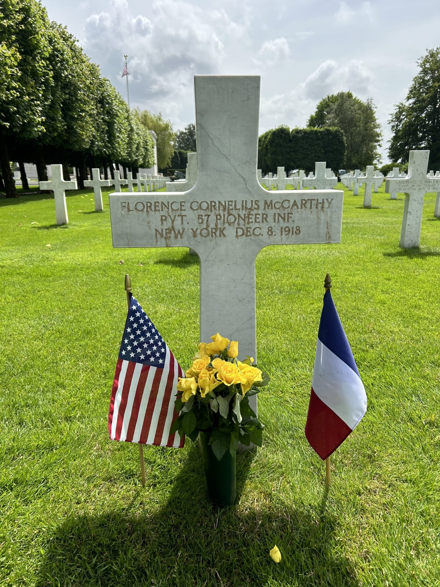 Croix blanche de la tombe de Florence Cornelius McCarthy frottée du sable des plages du débarquement. De part et d'autre de la croix se trouve un petit drapeau Américain et Français. Devant est planté un bouquet de roses jaunes.